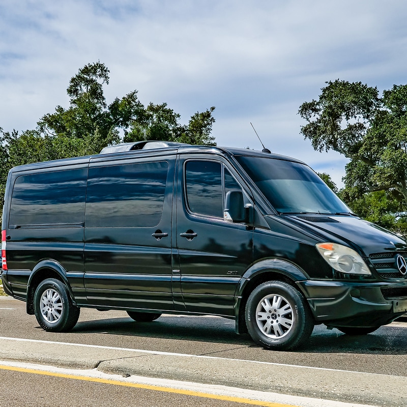 Van Mercedes Sprinter noir aux vitres teintées garé sur le bord d'une route sous un ciel nuageux.