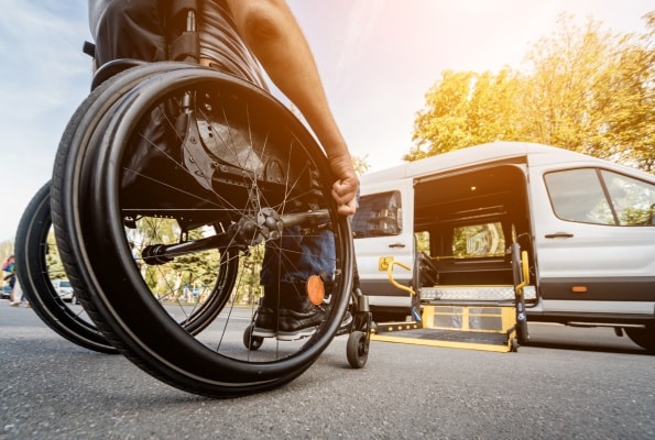 Transport PMR : Fourgon accessible avec rampe élévatrice Personne en fauteuil roulant vers un van PMR avec rampe élévatrice déployée par temps ensoleillé.