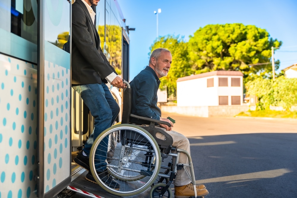 Accessibilité bus : aide d'un senior en fauteuil roulant Homme aidant un senior en fauteuil roulant à descendre d'un bus via une rampe d'accès sous le soleil.